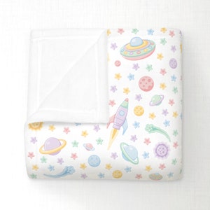May include: A white baby blanket with a colorful space-themed pattern. The design includes planets, stars, rockets, and UFOs in pastel colors. The blanket is folded, revealing a soft, white interior.