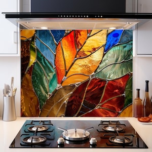 Può includere: Un paraschizzi da cucina colorato con un design a vetrata di foglie stilizzate nei toni del rosso, arancione, giallo, verde e blu. Il paraschizzi è installato sopra un piano cottura in vetro nero con quattro fuochi.