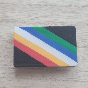 Puede incluir: Un broche rectangular que presenta una serie de rayas diagonales en negro, rojo, amarillo, blanco, azul y verde. El diseño está orientado de arriba a la izquierda hacia abajo a la derecha, creando un patrón colorido en este pequeño accesorio.