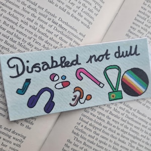 May include: A pale blue card with the handwritten text "Disabled not dull" and several colourful illustrations. These include a hearing aid, headphones, and a rainbow-striped circle.
