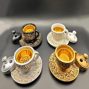 May include: Four ceramic incense burners shaped like coffee cups with saucers. Each burner has a gold-colored top with a decorative design and a lid. The burners are in black, white, and gold.