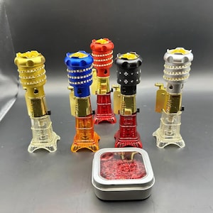 May include: Five different colored metal incense burners with a decorative design. Each burner has a round top with a small hole in the center and a base that resembles a tower. The burners are arranged in a row on a black surface. A small tin container with a clear lid filled with red incense is in the foreground.