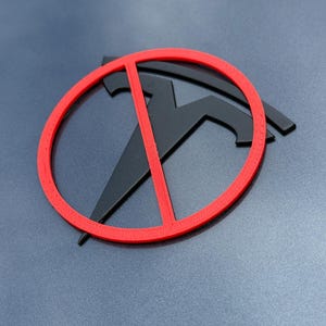 May include: A black Tesla logo with a red prohibition sign over it, set against a gray background. The design is a 3D-printed emblem, likely for a vehicle or similar application. The red circle and slash are prominent.