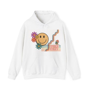 Può includere: Felpa con cappuccio bianca con una grafica in stile retrò che presenta un viso sorridente, fiori e il testo "Take it easy".