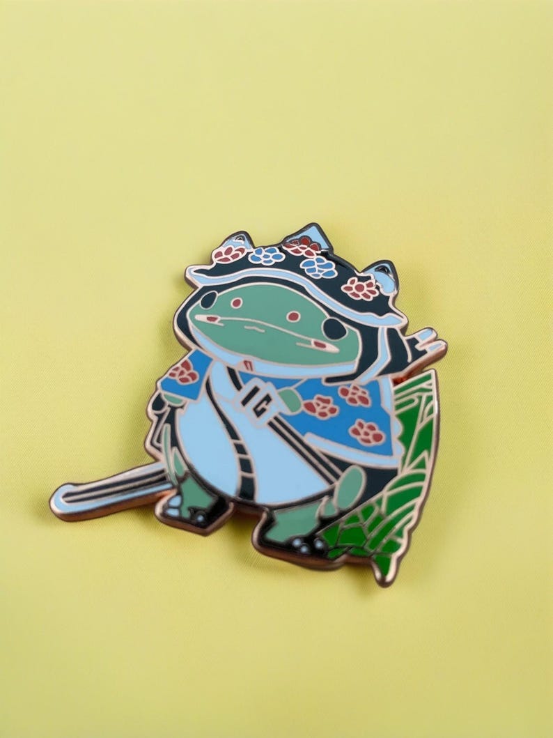 Peut inclure: Une grenouille verte portant un kimono bleu et blanc &agrave; motifs floraux et un chapeau de feuille verte. La grenouille tient une &eacute;p&eacute;e et porte une couronne de fleurs sur la t&ecirc;te.