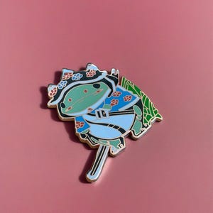 Peut inclure: Une grenouille verte portant un kimono bleu et blanc avec un motif de fleurs rouges et blanches. La grenouille tient une &eacute;p&eacute;e et a une feuille verte derri&egrave;re elle. La grenouille porte un chapeau noir avec des fleurs blanches.
