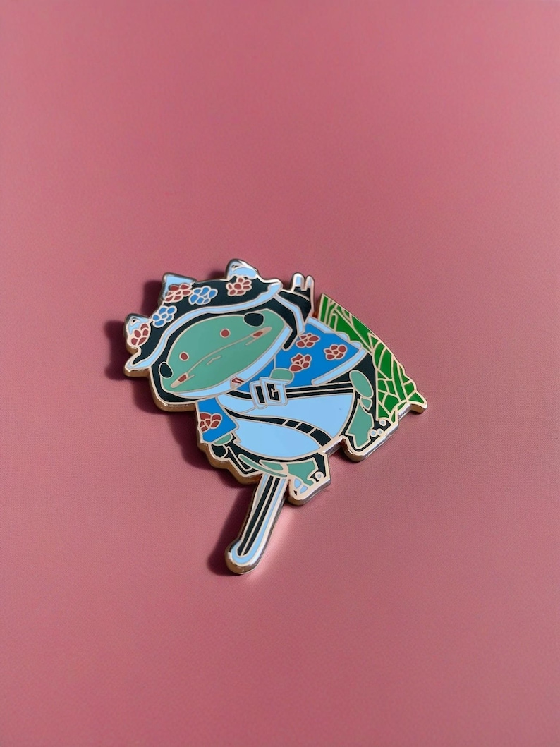 Peut inclure: Une grenouille verte portant un kimono bleu et blanc avec un motif de fleurs rouges et blanches. La grenouille tient une &eacute;p&eacute;e et a une feuille verte derri&egrave;re elle. La grenouille porte un chapeau noir avec des fleurs blanches.