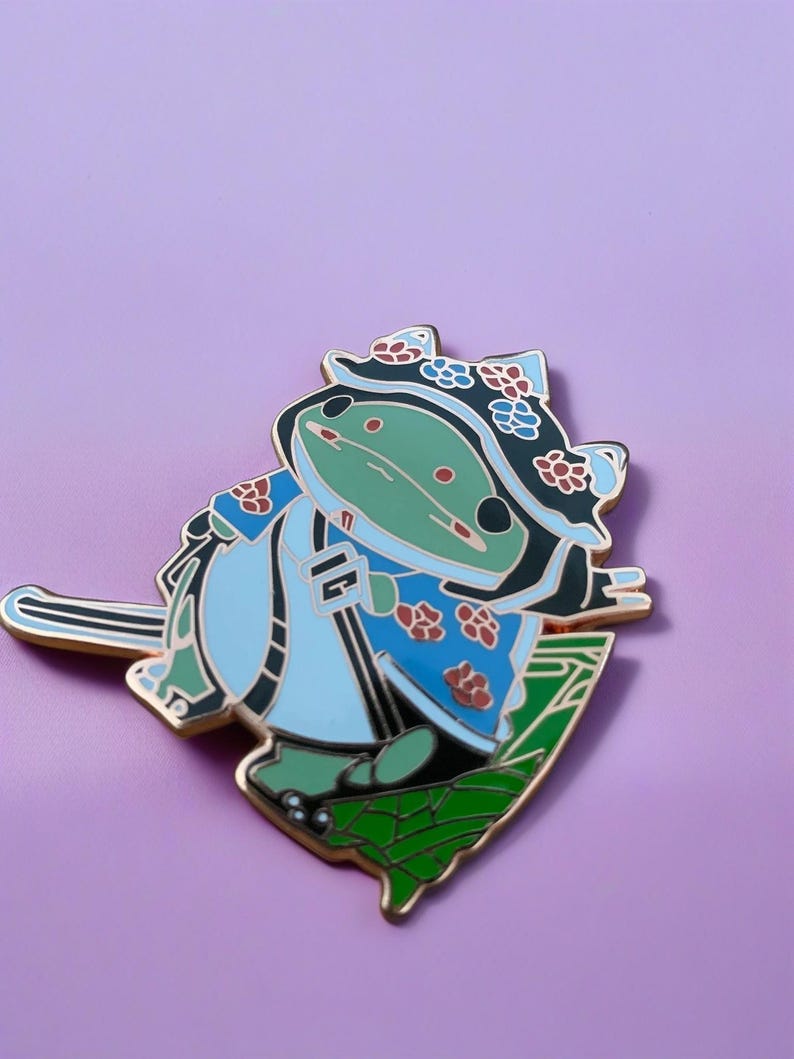 Peut inclure: Une grenouille verte portant un chapeau &agrave; fleurs bleu et blanc et une veste &agrave; fleurs bleu et blanc descend une colline verte en ski. La grenouille porte un masque de ski noir et tient un b&acirc;ton de ski dor&eacute; dans sa main.