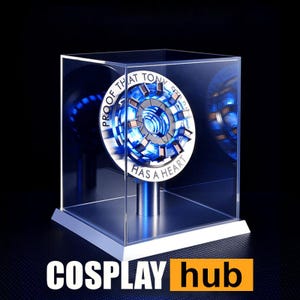 Może przedstawiać: Przezroczysta akrylowa gablota ze srebrną podstawą, prezentująca replikę reaktora łuku Tony'ego Starka wykonaną z niebieskiego i srebrnego metalu. Reaktor zawiera napis "Proof that Tony Stark has a heart".