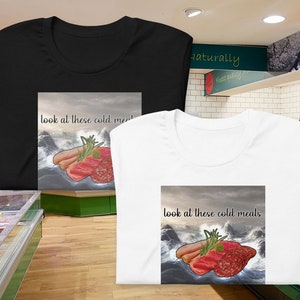 Può includere: Due t-shirt con un design grafico di una varietà di salumi su un mare tempestoso. Il testo sulle t-shirt recita "look at these cold meals".