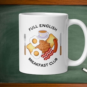 Può includere: Una tazza in ceramica bianca con un'illustrazione in bianco e nero di una colazione inglese completa, che include uova, pancetta, salsiccia, fagioli, toast e una tazza di tè. Il testo "Full English Breakfast Club" è stampato sulla tazza.