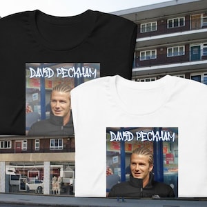 Può includere: Due t-shirt con una foto di David Beckham. La foto mostra Beckham con i capelli intrecciati e una giacca nera. Il testo "DAVID BECKHAM" è scritto in bianco su sfondo nero sopra la foto.
