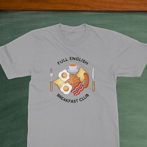 Può includere: T-shirt grigio chiaro con una grafica circolare raffigurante una colazione inglese completa: uova, toast, salsiccia, pancetta e caffè. Il testo "FULL ENGLISH BREAKFAST CLUB" è arcuato sopra e sotto l'illustrazione della colazione.