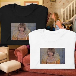 Può includere: Due t-shirt con un'immagine stampata di una donna che indossa un costume dorato e bianco con un grande colletto a pieghe. La donna guarda direttamente lo spettatore. Le t-shirt sono su un divano e uno sgabello.