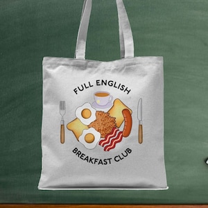 Può includere: Borsa tote bianca con una grafica di una colazione inglese completa, che include uova, pancetta, salsiccia, toast, fagioli e una tazza di tè. Il testo "Full English Breakfast Club" è stampato sulla borsa.