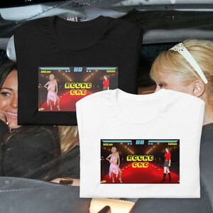 Può includere: Due magliette bianche con una stampa grafica di uno schermo di videogioco. Lo schermo mostra due donne con abiti diversi in piedi su un tappeto rosso. Il testo sullo schermo dice "Round One".