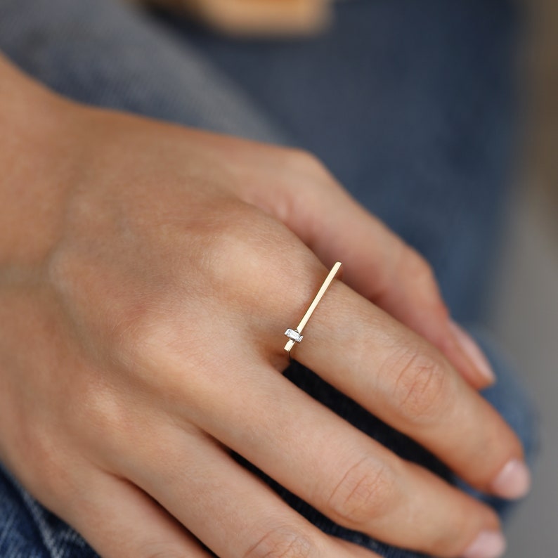 May include: A gold ring with a single, small, rectangular diamond set in the center of a thin, horizontal bar.