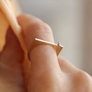 May include: A gold ring with a square band and a small diamond set in the top. The ring is worn on a person's finger.