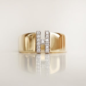 May include: A gold band ring with a diamond-encrusted letter 'H' in the centre.