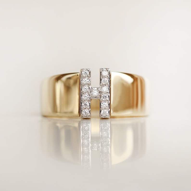 May include: A gold band ring with a diamond-encrusted letter 'H' in the center.
