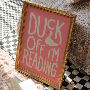 Könnte beinhalten: Ein rechteckiger Wandkunstdruck in einem goldenen, verzierten Rahmen mit rosa Hintergrund, weißem Text DUCK OFF I'M READING und einer kleinen weißen Entenillustration. Der Druck steht neben einem offenen Buch und einer Teetasse.