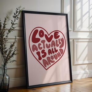 Pode incluir: Impressão artística emoldurada com fundo rosa e formato de coração grande. O coração contém as palavras "LOVE ACTUALLY IS ALL AROUND" em tons de vermelho. A impressão está em uma moldura preta e encostada em uma parede.