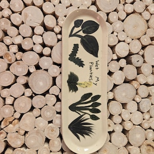 Puede incluir: Bandeja ovalada de cerámica color crema con ilustraciones de plantas negras y el texto manuscrito "Wet My Plantes". La bandeja tiene forma redondeada y está colocada sobre un fondo de rodajas de madera apiladas.