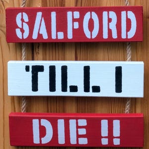 May include: Three wooden signs hanging on a rope. The top sign is red with white lettering that says "SALFORD". The middle sign is white with black lettering that says "TILL I". The bottom sign is red with white lettering that says "DIE!!".