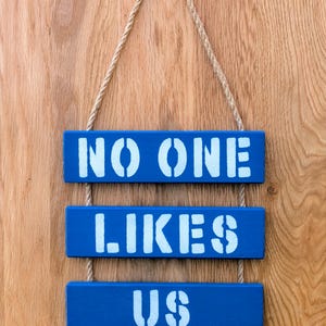 Pu&ograve; includere: Tre cartelli di legno blu appesi a una corda su uno sfondo di legno. I cartelli recitano "NO ONE", "LIKES" e "US" in vernice bianca.