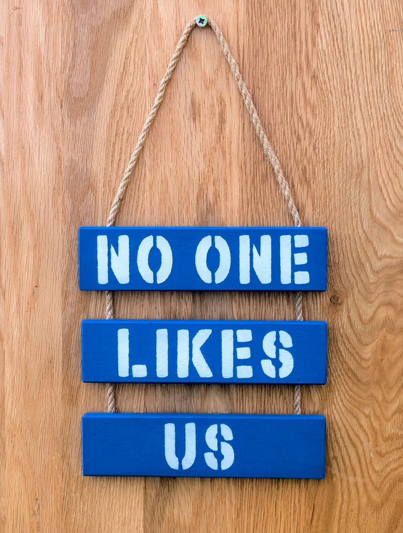 Pu&ograve; includere: Tre cartelli di legno blu appesi a una corda su uno sfondo di legno. I cartelli recitano "NO ONE", "LIKES" e "US" in vernice bianca.