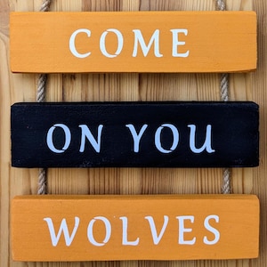 May include: Three wooden signs hanging from twine, painted in orange and black. The top sign reads "COME", the middle sign reads "ON YOU", and the bottom sign reads "WOLVES".