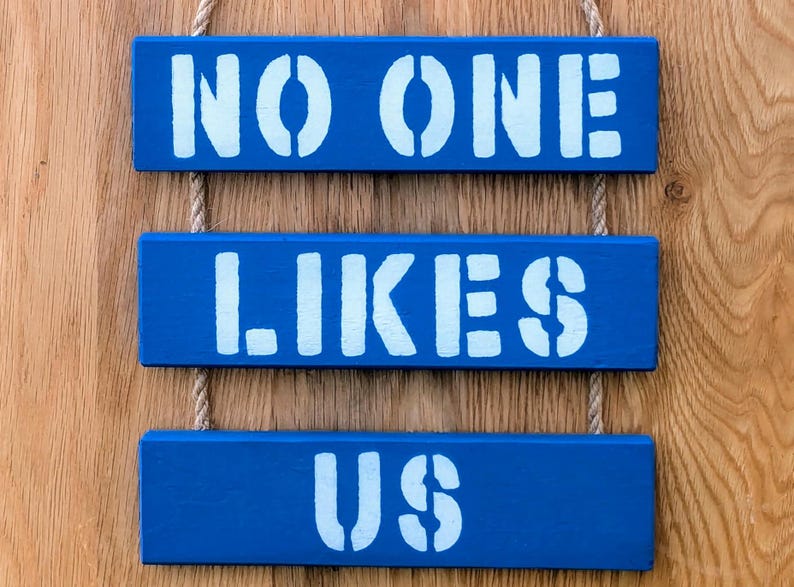 Pu&ograve; includere: Tre cartelli in legno blu con testo bianco dipinto appesi a uno sfondo in legno. I cartelli recitano "NO ONE", "LIKES" e "US".