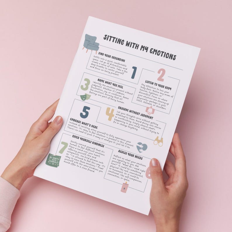 May include: A white paper titled "Sitting With My Emotions" is held by hands. The paper features text and numbered steps for emotional well-being, with illustrations of a chair and other icons. The text includes phrases like "Find Your Grounding" and "Listen to Your Body".