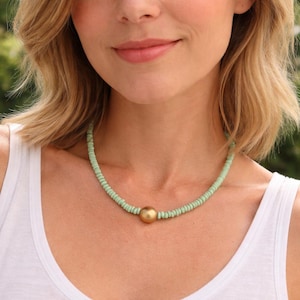 May include: A delicate necklace featuring small, light green beads and a central gold-colored sphere. The necklace is worn around the neck, creating a simple yet elegant accessory. The necklace is a stylish piece of jewelry.