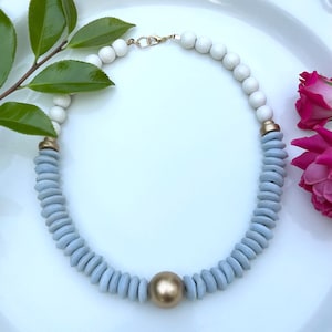 May include: A necklace with a gold clasp and a gold sphere pendant. The necklace is made of white and light blue wooden beads. The necklace is displayed on a white surface with green leaves and pink flowers.