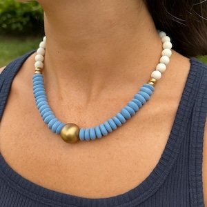 May include: A statement necklace featuring a large gold-colored sphere pendant and a mix of blue and white wooden beads. The necklace has gold-colored metal accents and is worn on a person wearing a navy blue top.