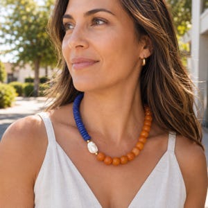 May include: A beaded necklace featuring a section of small, cylindrical blue beads and a section of larger, round orange beads. A single, irregular white pearl is positioned between the two bead types, accented by small gold-toned metal spacers.