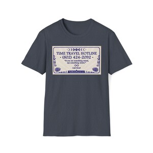 May include: A navy blue t-shirt featuring a rectangular graphic with the text "TIME TRAVEL HOTLINE (802) 424-2092" and the phrase "If you see something weird, say something weird." The graphic has a vintage design with decorative elements.