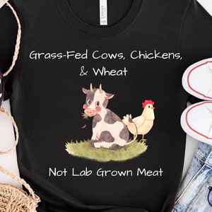 May include: Black t-shirt with a white graphic of a cow, chicken, and wheat. The text "Grass-Fed Cows, Chickens, & Wheat" is above the graphic and "Not Lab Grown Meat" is below the graphic.