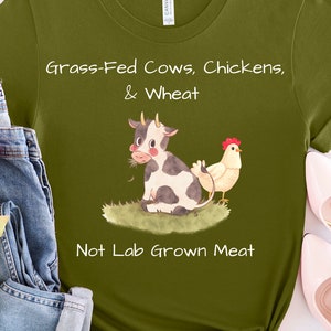 May include: Green t-shirt with a white graphic of a cow and a chicken. The text on the shirt reads "Grass-Fed Cows, Chickens, & Wheat Not Lab Grown Meat".