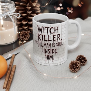 Pu&ograve; includere: Tazza in ceramica bianca con la scritta "WITCH KILLER, THE HUMAN IS STILL INSIDE HIM" in un font a goccia simile al sangue. La tazza ha un design a muro di mattoni ed &egrave; riempita con un liquido scuro.