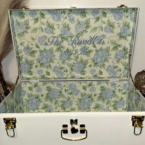 May include: White decorative chest with gold hardware and a floral interior. The inside lid features a light green and white floral pattern with blue hydrangea flowers and the text "The Russell's 06.13.26" in blue script.