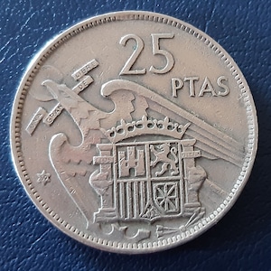 以下が含まれることがあります： 25 PTASの価値を持つスペインの銀貨。コインには、城とライオンが描かれた紋章があり、翼のある鷲に囲まれています。コインの左側には「UNA GRAN LIBRE DE」という文字が見えます。