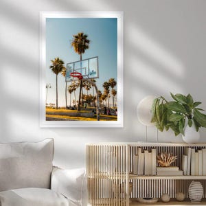 May include: A framed print featuring a basketball hoop with a red net, set against a backdrop of palm trees and a clear blue sky. The image captures a sunny day at a beachside court, with the word "CLIPPERS" visible on the backboard.