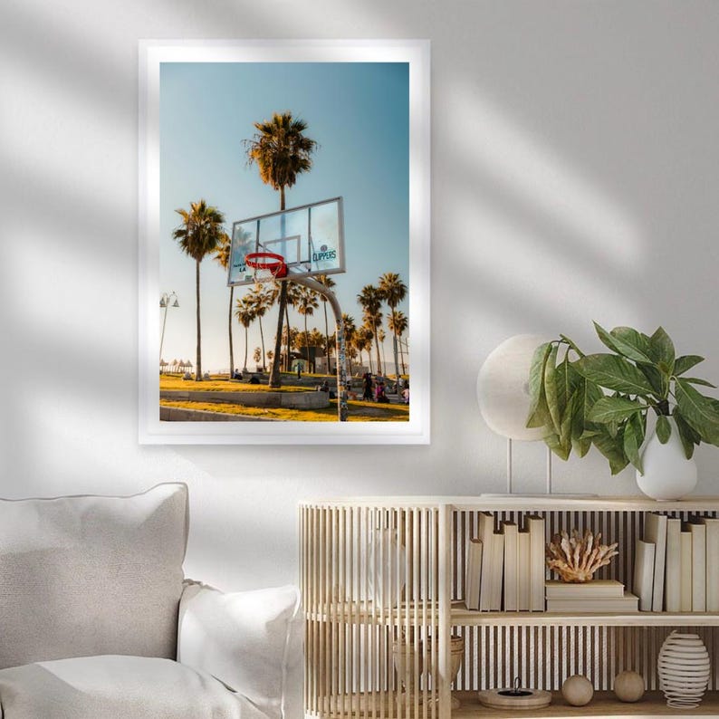 May include: A framed print featuring a basketball hoop with a red net, set against a backdrop of palm trees and a clear blue sky. The image captures a sunny day at a beachside court, with the word "CLIPPERS" visible on the backboard.
