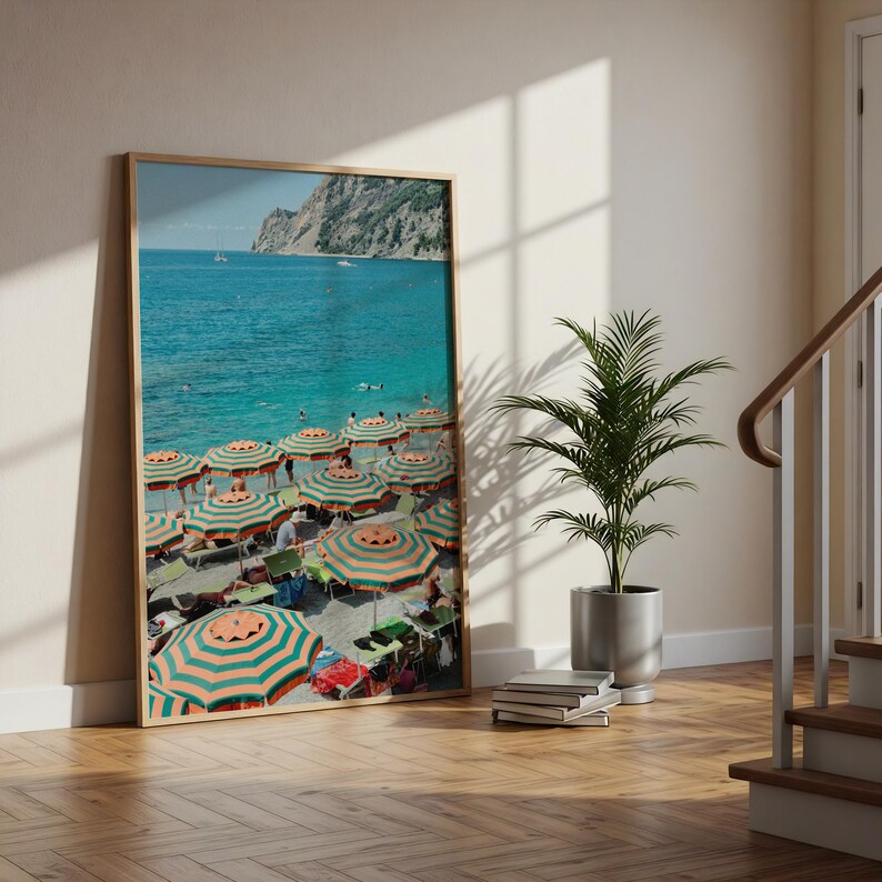 May include: A framed print depicts a vibrant beach scene. The image showcases a turquoise sea, a rocky coastline, and numerous beach umbrellas with orange and green stripes. People are visible on the beach, and sailboats dot the horizon.