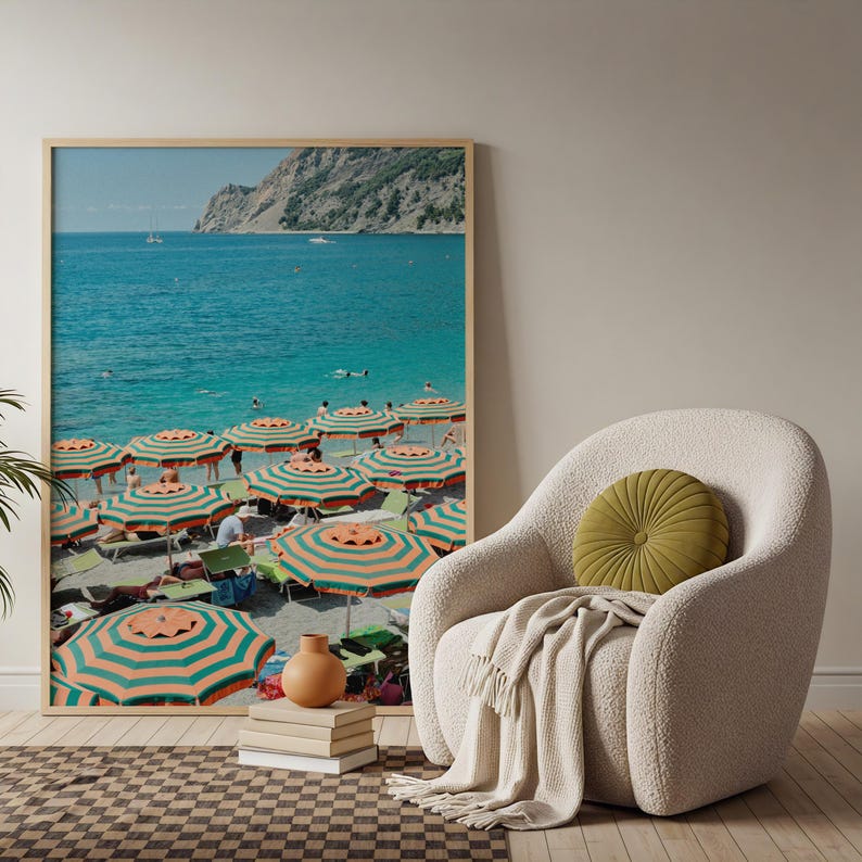 May include: A framed print of a beach scene with turquoise water and striped orange and green umbrellas. The print is displayed next to a cream-colored armchair with a green cushion and a beige throw blanket.
