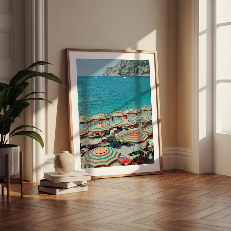 May include: A framed print of a beach scene with turquoise water and a sandy shore. Numerous striped orange and green umbrellas dot the beach, providing shade. A mountain range is visible in the background. The print is displayed on a wooden floor.