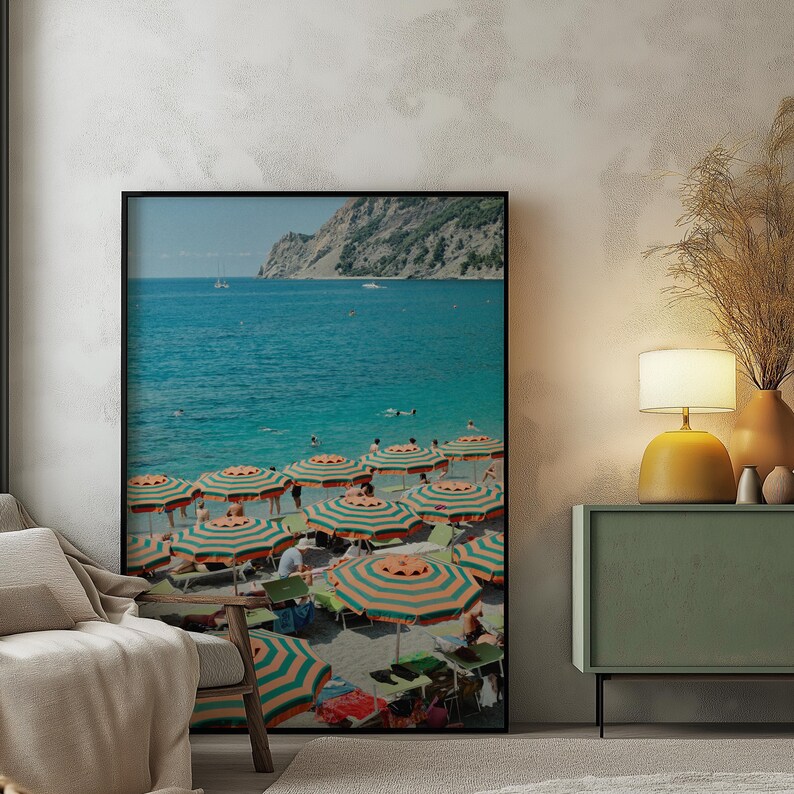 May include: A framed print of a beach scene with turquoise water and a mountain in the background. Striped orange and green umbrellas dot the sandy shore, with people relaxing underneath. The image evokes a sunny, seaside vacation.