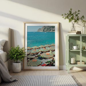 May include: A framed print of a beach scene with striped orange and green umbrellas. The turquoise water and a rocky cliff are visible in the background. The print is displayed in a light-colored wooden frame.